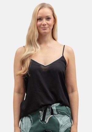 Donna bionda con capelli lunghi che indossa un top nero con spalline sottili e pantaloni verdi con motivi, in piedi davanti a uno sfondo chiaro e semplice.