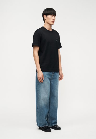 Άνδρας που στέκεται φορώντας μαύρο κοντομάνικο μπλουζάκι, φαρδιά μπλε τζιν και μαύρα παπούτσια, μπροστά σε απλό λευκό φόντο.