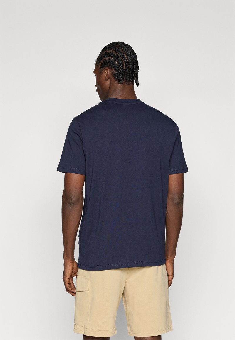 T-shirt en coton bleu marine avec un col rond, des manches courtes et une coupe décontractée. Texture lisse et coutures minimales ; porté avec un short beige.