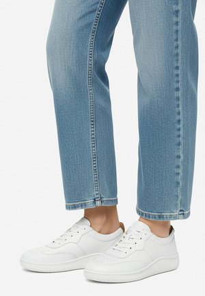 Person, die hellblaue Jeans mit Umschlag und weiße Leder-Schnürsneaker trägt und vor weißem Hintergrund steht.