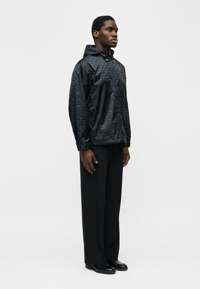 Άνδρας που φοράει μαύρο εμπριμέ μπουφάν με κουκούλα, μαύρο παντελόνι με φαρδιά πόδια και μαύρα δερμάτινα παπούτσια, στέκεται μπροστά σε ένα απλό λευκό φόντο.