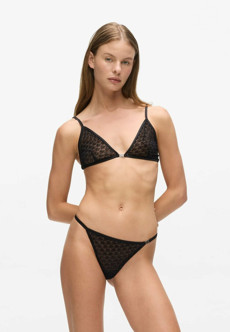 Femme portant un soutien-gorge triangle en dentelle noire et un string assorti, posant avec les mains dans le dos sur fond blanc.