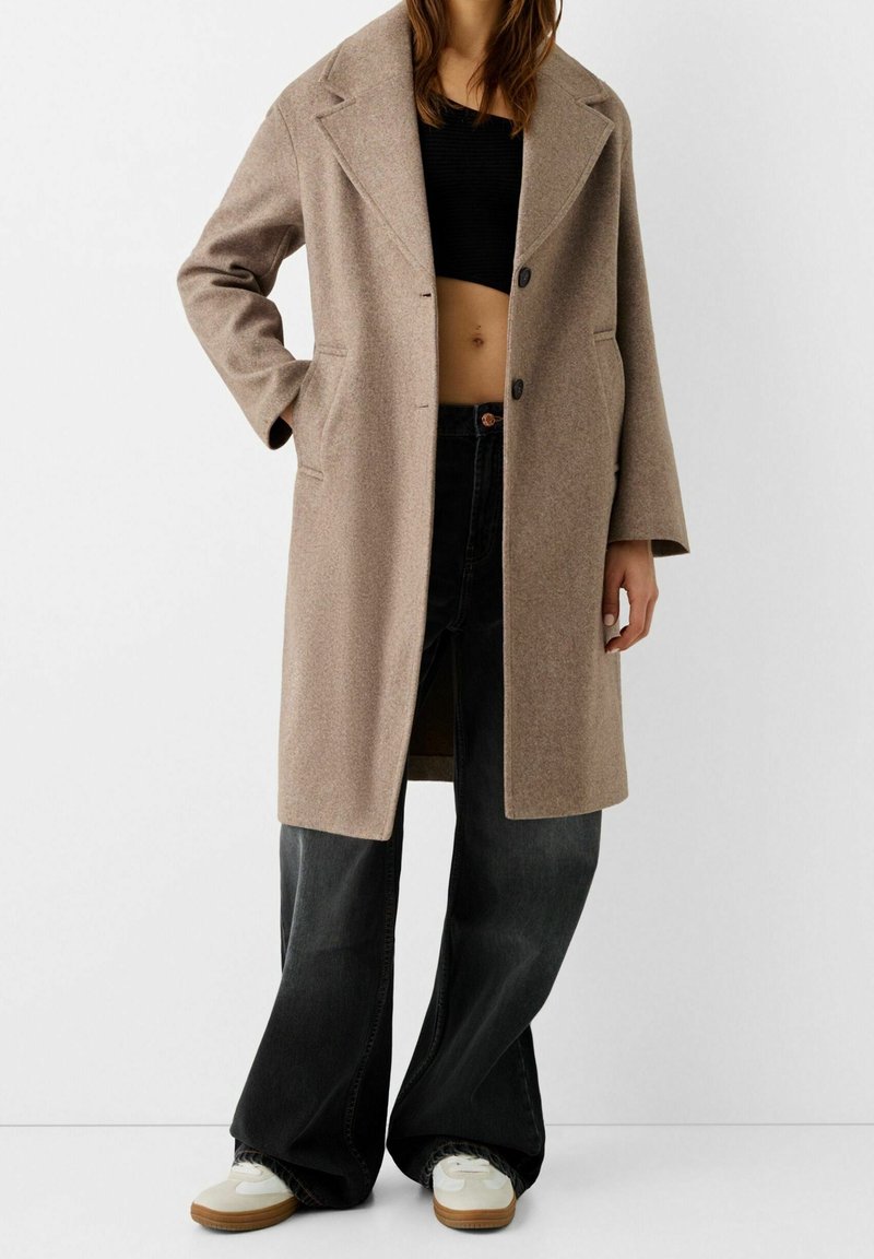 Cappotto lungo in misto lana marrone con ampie revers, chiusura con un bottone e tasche laterali, indossato sopra un top cropped nero e jeans larghi scuri.