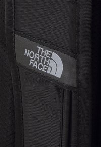 Czarna łatka z tkaniny z białym logo "THE NORTH FACE", z zaokrąglonym wzorem i teksturowaną powierzchnią, wszyta w materiał o tym samym kolorze.
