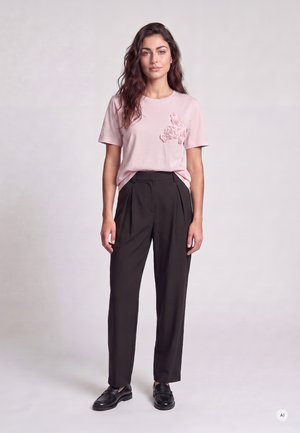 Junge Frau mit langen welligen Haaren, die ein hellrosa T-Shirt mit Blumendetail, schwarze hoch taillierte Hose und schwarze Loafers trägt, stehend.