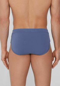 Pánské plavkové slipy, jednobarevný světle modrý materiál, hladká textura, střední pas, minimalistický design, bez viditelného kování nebo vzorů.