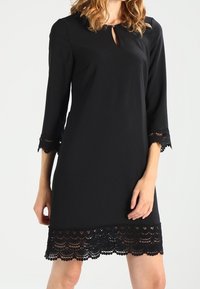 Kvinna i en svart knälång klänning med 3/4-långa ärmar och spetsdetaljer på muddar och nederkant, stående framför en neutral bakgrund.