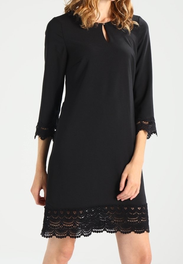 Kvinna i en svart knälång klänning med 3/4-långa ärmar och spetsdetaljer på muddar och nederkant, stående framför en neutral bakgrund.