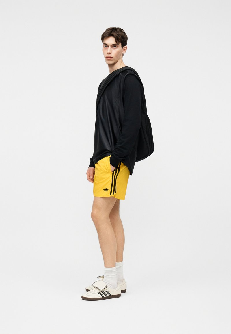 Ung mand står i profil iført sort langærmet trøje, gule Adidas-shorts, hvide sokker, hvide sneakers og bærer en sort taske over den ene skulder.