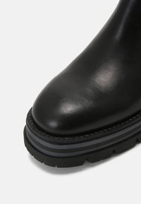 Botte en cuir noir avec embout arrondi et semelle épaisse à crampons superposés en noir et gris foncé sur fond blanc.