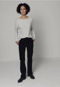 Hellgraues Langarmshirt in lockerer Passform, kombiniert mit schwarzen, eng anliegenden Jeans. Das Modell trägt schwarze Schuhe und steht vor einem neutralen Hintergrund.