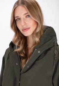 Olijfgroene hoodiejas met een gestructureerde afwerking, voorzien van een volledige rits, drukknoopsluiting en trekkoordaccenten rond de capuchon.