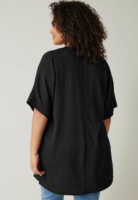 Camicia nera oversize in tessuto leggero, con maniche corte, una cucitura posteriore e orlo arrotondato. Indossata con jeans blu.