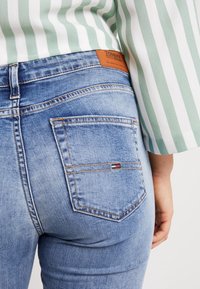 Närbild av en person som bär ljusblå jeans med en Tommy Jeans-tag på linningen och sträcker sig efter bakfickan. Rutig skjorta synlig.