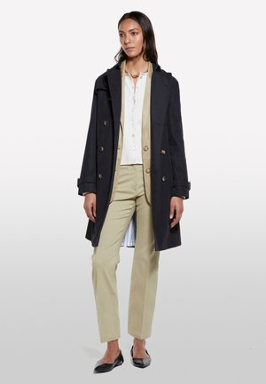 Cappotto lungo blu navy con bottoni doppi, abbinato a una camicia e pantaloni di un beige chiaro. La modella indossa delle scarpe nere flat, visibile la trama del tessuto.