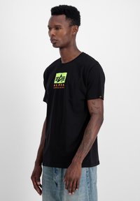 Must värvitud puuvillane T-särk, millel on roheline ruudukujuline logo, kus on kirjas "ALPHA" oranži tekstiga. Lühikesed varrukad ja klassikaline lõige, millel on sile tekstuur.