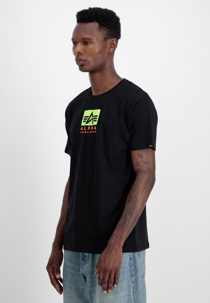 Must värvitud puuvillane T-särk, millel on roheline ruudukujuline logo, kus on kirjas "ALPHA" oranži tekstiga. Lühikesed varrukad ja klassikaline lõige, millel on sile tekstuur.