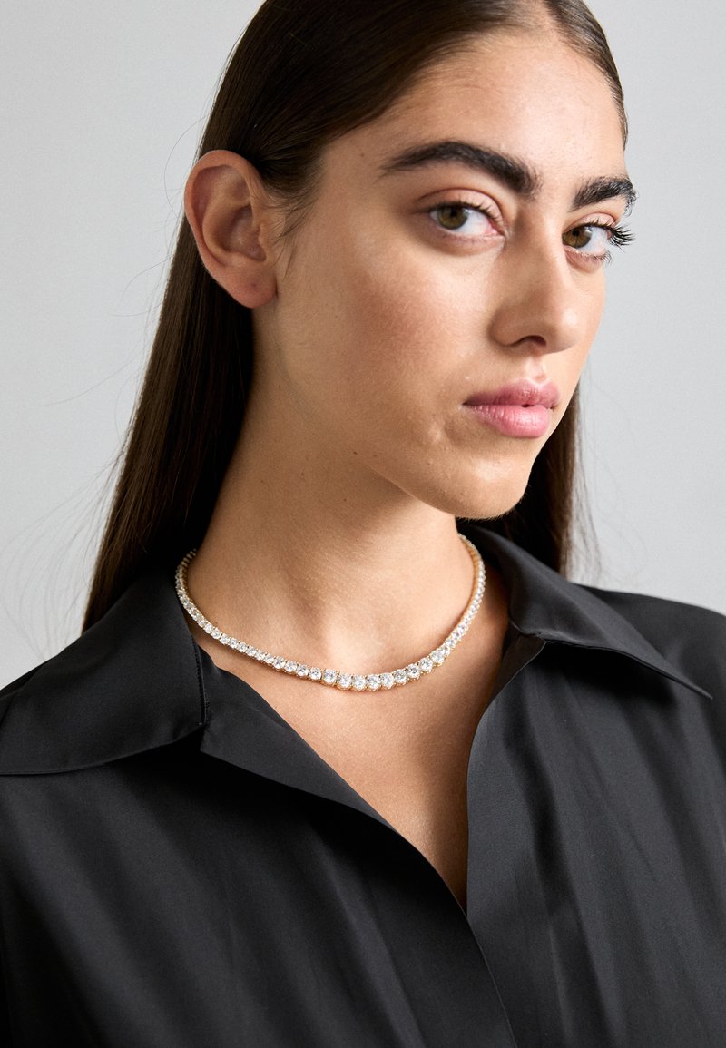 Collana in cristallo trasparente con montatura dorata, caratterizzata da un design curvo con cristalli di varie dimensioni, indossata sopra una camicia nera con colletto.