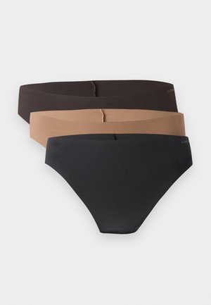 Ung kvinde stående barfodet mod en ensfarvet baggrund iført en sort bh og matchende sort undertøj, poserer med en afslappet hånd.