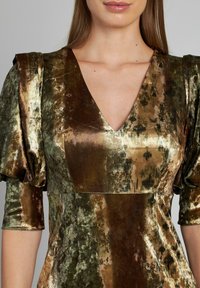 Donna che indossa un abito in tessuto lucido e testurizzato con scollo a V e maniche a metà gonfiate, in tonalità metalliche di verde, bronzo e oro.