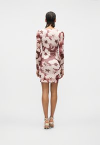 Γυναίκα με πλεξούδες, φορώντας εφαρμοστό μακρυμάνικο φόρεμα καφέ και κρεμ με λουλούδια και μπεζ πέδιλα με τακούνι, στέκεται μπροστά σε λευκό φόντο.