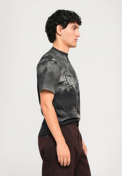 Young man with curly hair wearing a short-sleeve black and gray graphic t-shirt and dark brown pants facing right in a neutral background.