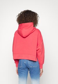 Korálová zkrácená mikina s kapucí na stáhnutí, širokými rukávy a žebrovanými manžetami, spárovaná s džínami v světle modrém gradientu.