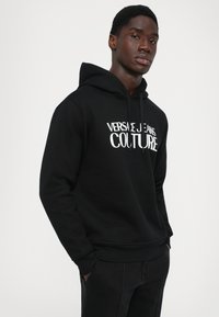 Νεαρός άνδρας που φορά μαύρη κουκούλα με το λογότυπο "Versace Jeans Couture", που στέκεται με τα χέρια στις τσέπες του σε λευκό φόντο.