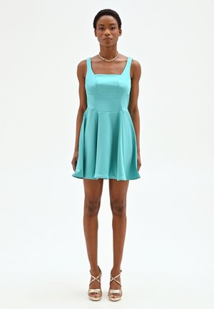 Femme debout, portant une robe turquoise sans manches avec un décolleté carré et des talons hauts dorés à lanières, fond neutre.