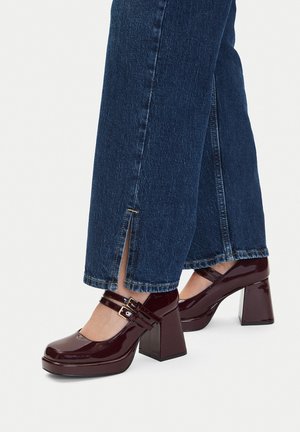 Bordowe lakierowane platformy na grubym obcasie z podwójnymi paskami na kostce, zaokrąglonym noskiem i wyraźnie podwyższonym obcasem. Noszone z ciemnoniebieskimi dżinsami.