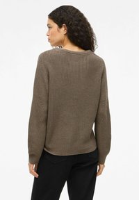Femme aux cheveux bouclés portant un pull tricoté marron et un pantalon noir, de profil contre un fond blanc uni.