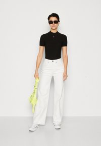 Body à manches courtes noir ajusté, jeans blancs à jambes larges, sac transparent avec des anses jaune fluo, baskets blanches et lunettes de soleil noires.
