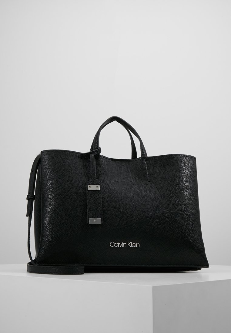 Czarna skórzana torba na ramię o teksturowanym wykończeniu, z podwójnymi uchwytami, odpinanym paskiem, srebrnym okuciem oraz wytłoczonym logo "Calvin Klein" z przodu.