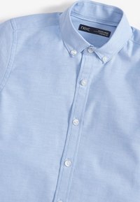 Chemise bleu clair à boutons sur le devant avec col, montrant l’étiquette de la marque « next » à l’intérieur de la zone du cou.