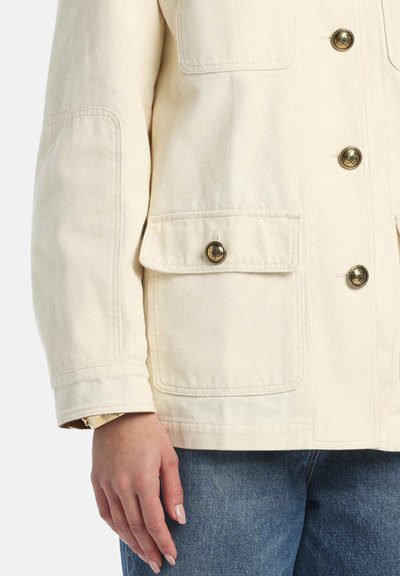 Cremefarbene Canvasjacke mit goldfarbenen Knöpfen, zwei Vorderaschen und leicht oversized Passform, kombiniert mit hellblauen Jeans.