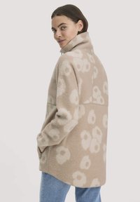 Vrouw in een beige fleece jack met witte bloemenpatroon en hoge kraag, handen in de zakken, gecombineerd met blauwe jeans, kijkt over haar schouder.
