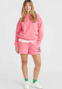 Felpa rosa con cappuccio e disegno testuale, abbinata a pantaloni corti rosa con stampa nera. Completata con calzini verdi e sneakers bianche.