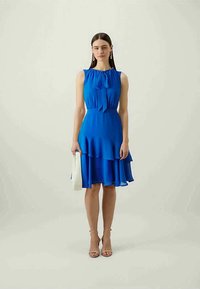 Modrá bezrukávová šaty s detailom mašle na výstrihu, vrstvené volány na leme, v kombinácii so svetlými sandálmi na opätku a kabelkou.