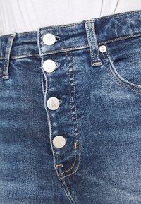 Jeans de mezclilla azul oscuro con una cintura alta, cinco botones con logotipos grabados y costuras en contraste a lo largo de las costuras.