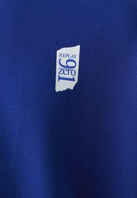 Tessuto blu con una superficie liscia e leggermente strutturata, presenta un'etichetta bianca con la scritta "REPLAY zero 91" in un carattere audace.
