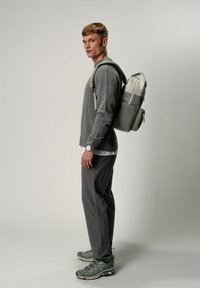 Zaino grigio realizzato in materiale sintetico, con spallacci regolabili, forma aerodinamica e chiusura con zip. Il modello indossa una maglietta grigia, pantaloni e scarpe verdi.