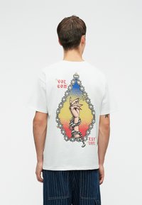 T-shirt blanc en coton avec un design graphique représentant une main, des chaînes, et un fond dégradé coloré. Impressions sur l'étiquette "VOLCOM" et "EST 1991".