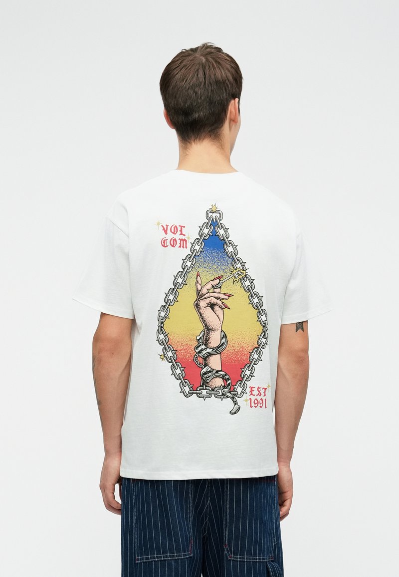 T-shirt blanc en coton avec un design graphique représentant une main, des chaînes, et un fond dégradé coloré. Impressions sur l'étiquette "VOLCOM" et "EST 1991".