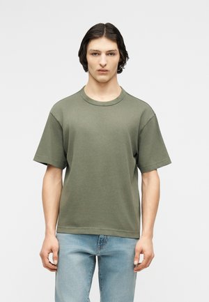 Ung person med skulderlangt mørkt hår iført en ensfarvet olivengrøn t-shirt med korte ærmer og lyseblå jeans mod en hvid baggrund.