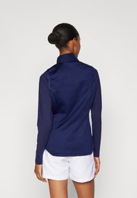 Mornarska dugosleeve majica s patentnim zatvaračem i strukturiranim ovratnikom, glatka tkanina, sa šivenim logotipom na leđima, u kombinaciji s bijelim šortsem.