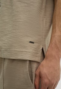 Beige strukturierte Bluse mit leichtem Glanz. Besitzt einen seitlichen Schlitz und ein kleines schwarzes "JOOP!"-Etikett am Saum. Glattes, gewebtes Material.