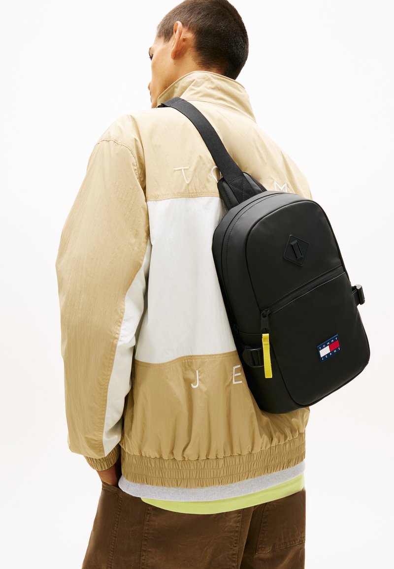 Μαύρο σακίδιο με λεία υφή, διαθέτει εμφανή κίτρινη τσέπη με φερμουάρ και μια λογότυπο σε κόκκινο, άσπρο και μπλε στο μπροστινό μέρος.