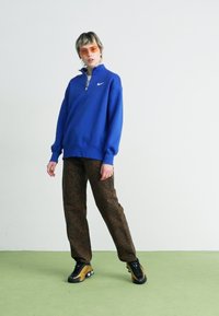 Sudadera azul con cierre de cuarto, que presenta un pequeño logo. Pantalones marrones con estampado y zapatillas negras con un acento amarillo. Gafas de sol naranjas.