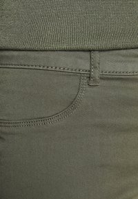 Olivgröna jeans har ett mjukt, stretchigt tyg med en subtil glans, överkantade fickor och kontrasterande sömmar längs sömmarna.