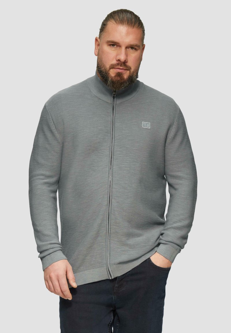 Grauer Reißverschluss-Pullover mit geripptem Material und Stehkragen, präsentiert mit einem kleinen Logodetail auf der Brust. Kombiniert mit dunklen Hosen.